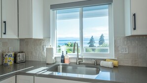 A kitchen with a large window, white cabinets, a stainless steel sink, and a view of the outdoors. - Water & Mountain Views: Escape to Whidbey Island! (Freeland)