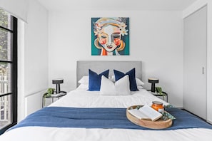 A modern bedroom with a large bed, two bedside tables, a window, and a painting on the wall.