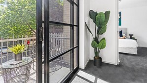 Une chambre d’hôtel moderne dotée d’une grande fenêtre, d’un balcon avec une petite table et un pot de fleurs, et d’un lit avec une tête de lit blanche.