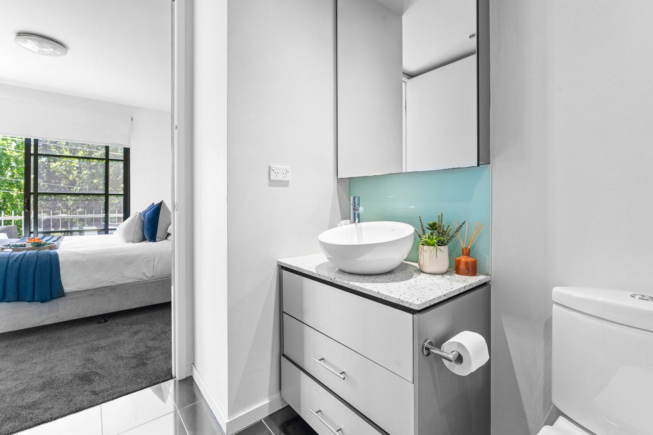 A modern bathroom with a white sink, a mirror, and a toilet. There is a window with a view of trees and a bed with a blue blanket in the adjacent room.
