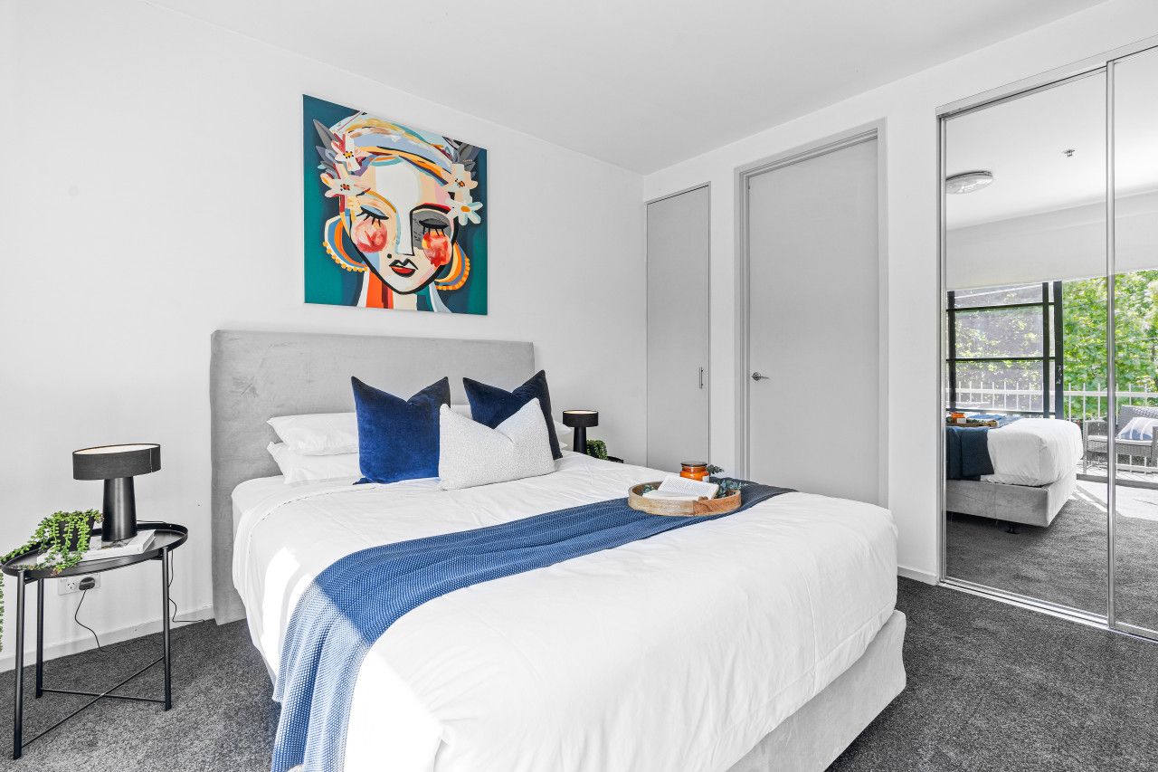 A modern bedroom with a large bed, a bedside table, a nightstand, a painting, and a sliding glass door leading to a balcony.