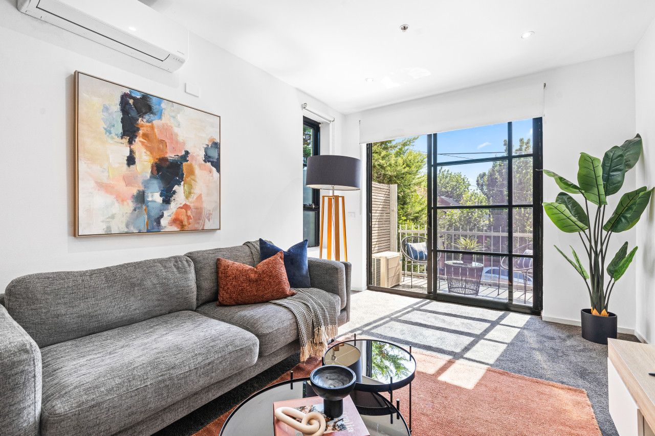 A modern living room with a grey sofa, a large abstract painting, a floor lamp, and a view of a balcony with outdoor furniture.