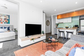 A modern living room with a flat-screen TV, a sofa, a coffee table, and a kitchen area with a bar stool.