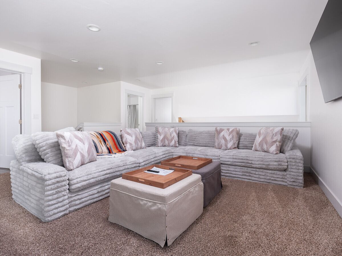 A modern living room with a grey sectional sofa, two ottomans, and a flat-screen TV mounted on the wall.