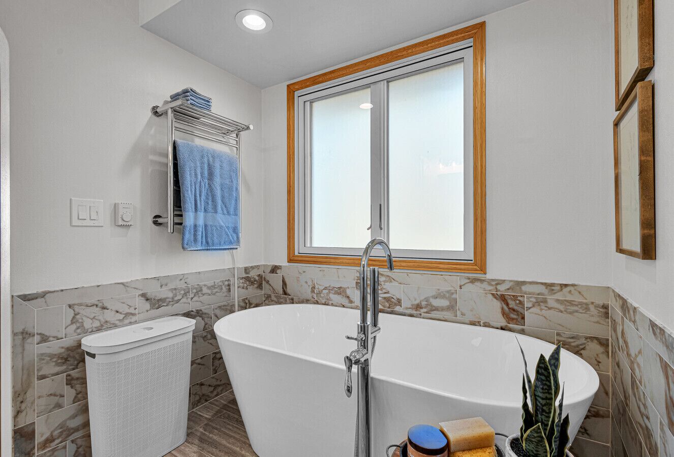 Un baño moderno con una bañera independiente, un perchero con una toalla azul, un cesto de basura y una ventana con marco de madera.