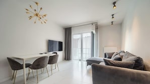 Ein modernes Wohnzimmer mit Fliesenboden, einem grauen Sofa, einem Esstisch mit vier Stühlen und einem an der Wand befestigten Fernseher.