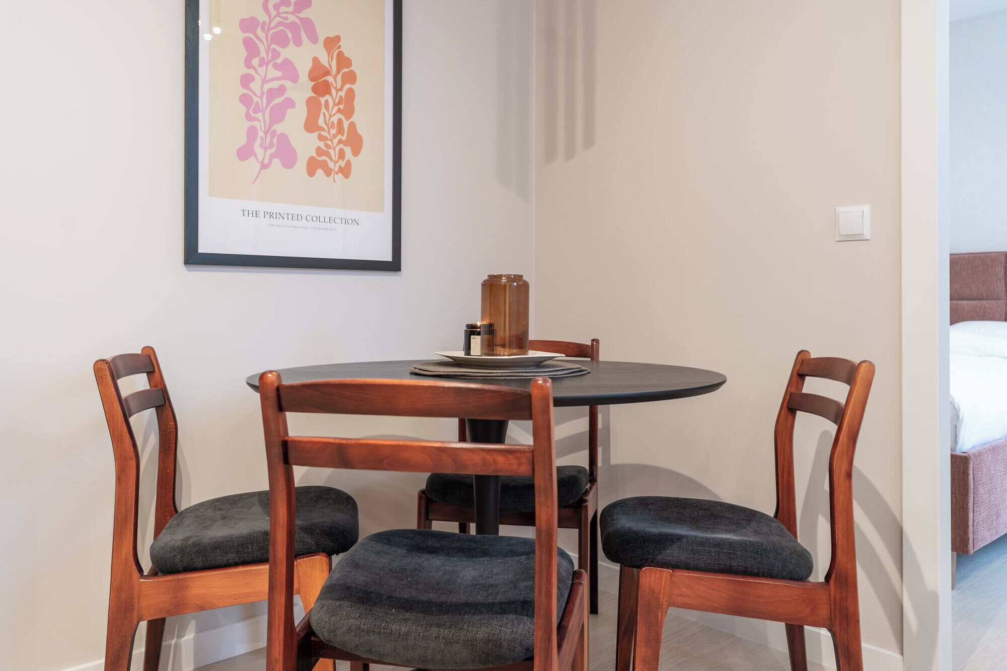 Un coin repas avec une table ronde, quatre chaises en bois, une œuvre d’art encadrée au-dessus de la table et un lit en arrière-plan.