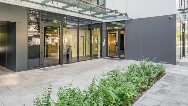 Un edificio moderno con entrada de vidrio, un paseo techado y una zona ajardinada con plantas.
