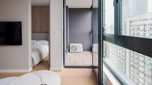 Un dormitorio moderno con cama, televisor y vistas a la ciudad a través de amplias ventanas.