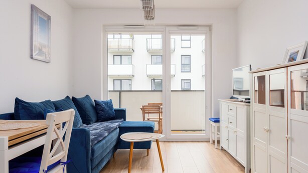 Una sala moderna con un sofá azul, una mesa de comedor de madera y una mesita redonda. Hay un armario blanco con un televisor encima.
