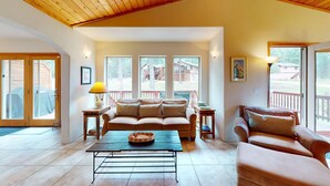 A spacious living room with a large sofa, a coffee table, and a second seating area with a cushioned bench. There are multiple windows providing natural light and views of the outside. - Convenient Location | Fireplace | Pet Friendly | Country Club Villas 13 (Angel Fire)