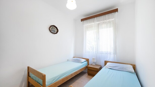 A small, clean room with two wooden beds, a bedside table with a lamp, and a window with sheer curtains. - 4 bedroom amazing apartment in Pirovac (Pirovac)