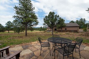An outdoor patio with a round table and chairs, surrounded by stone flooring, with a house and trees in the background. - Stunning Cimarron Golf Villa                           Lic# STR-1-2024-038 (Ridgway)