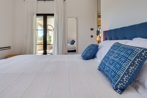 Ein Schlafzimmer mit einem großen Bett, weißer Bettwäsche, einem blau gemusterten Kissen und Blick durch eine Glastür in einen Außenbereich.