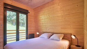 A wooden cabin bedroom with a bed, two bedside lamps, a nightstand, and a sliding glass door leading to an outdoor area. - Chalet du Souillet with Mountain Views, Private Terrace and Wi-Fi (Saint-Pierre-d'Entremont)