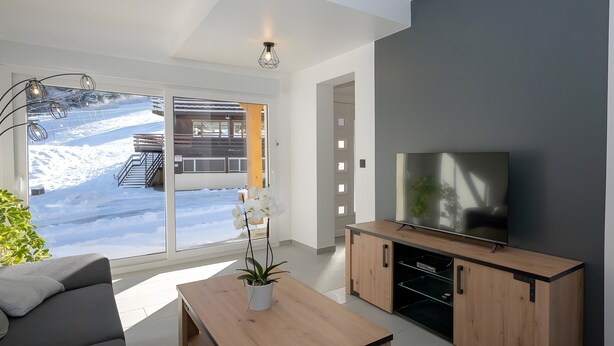 A modern living room with a sofa, a wooden coffee table, a TV, and a sliding glass door leading to a balcony with a view of a building and snow. - Chalet du Souillet with Mountain Views, Private Terrace and Wi-Fi (Saint-Pierre-d'Entremont)
