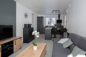 En moderne stue med et fladskærms-tv, et træsofabord med en plante, en grå sofa med puder, og et køkkenområde i baggrunden.