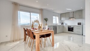 A modern kitchen with white cabinets, a dining area with a wooden table and chairs, and large windows offering a view. - Villa 'Costacabana -  La Luna' with Sea View, Private Pool and Wi-Fi (Lloret de Mar)