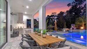 A modern outdoor dining area with a pool and a view of the sunset. - Luxe Retreat | 9BR Luxe Villa w/ Saltwater Pool, Gym, Theater, and Arcade Near Orlando Theme Parks (Kissimmee)