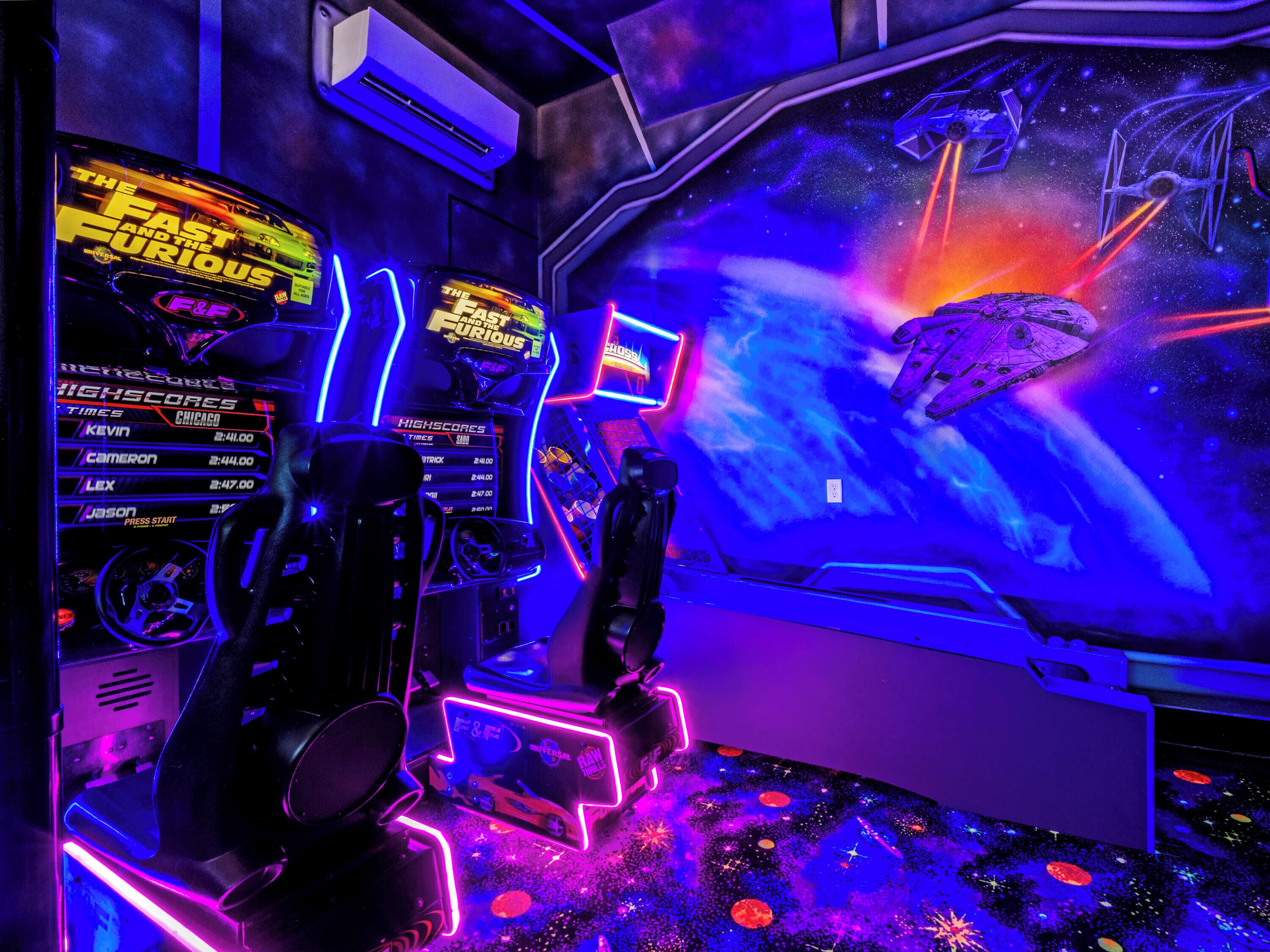 Un área de juegos con luces de neón, una configuración para juego de carreras y un mural con temática espacial.