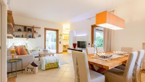 A modern living room with a dining area, featuring a wooden dining table, white chairs, and a large window.
