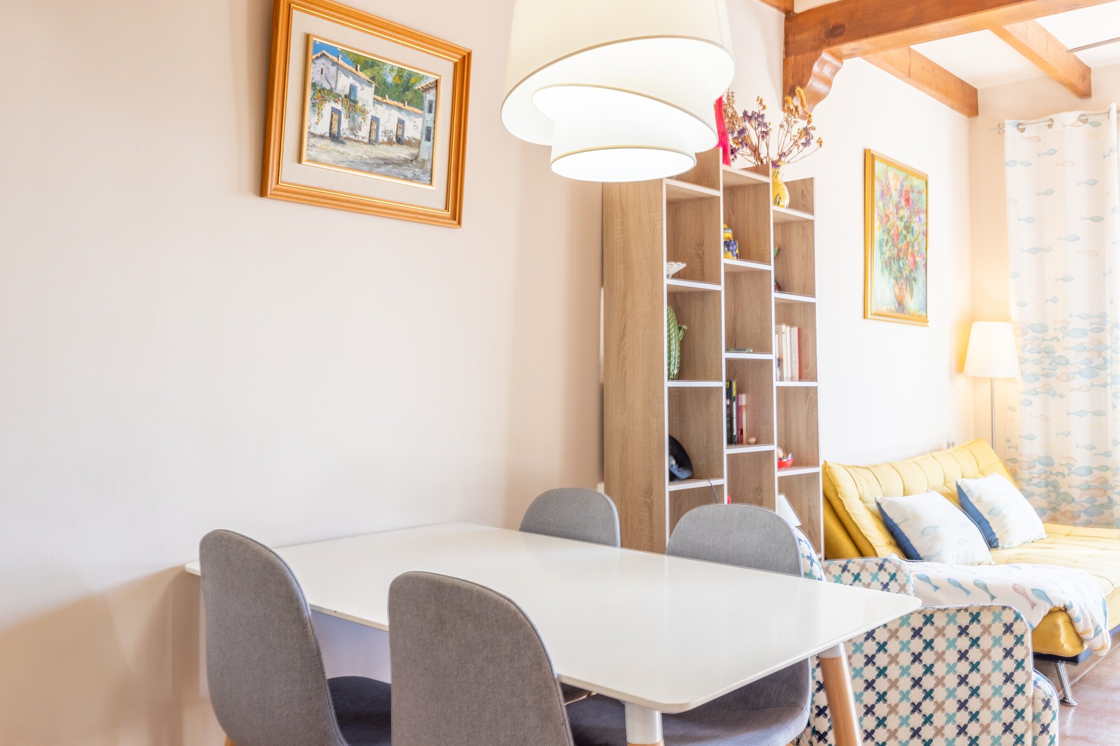 A room with a dining table, chairs, a bookshelf, a bed with patterned bedding, and a ceiling light fixture.