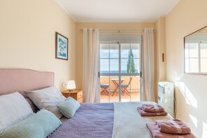 A bedroom with a bed, pillows, a bedside table with a lamp, a window with curtains, and a view of a balcony with a table and chairs.