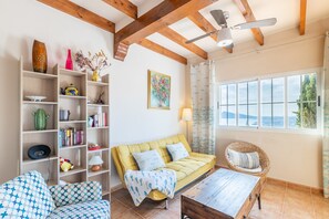 Un salon avec un canapé jaune, un fauteuil en osier, une table basse en bois et une bibliothèque contenant divers objets.