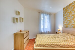 Une chambre à coucher avec un lit, une commode en bois, une table de chevet, une lampe et une fenêtre avec des rideaux.