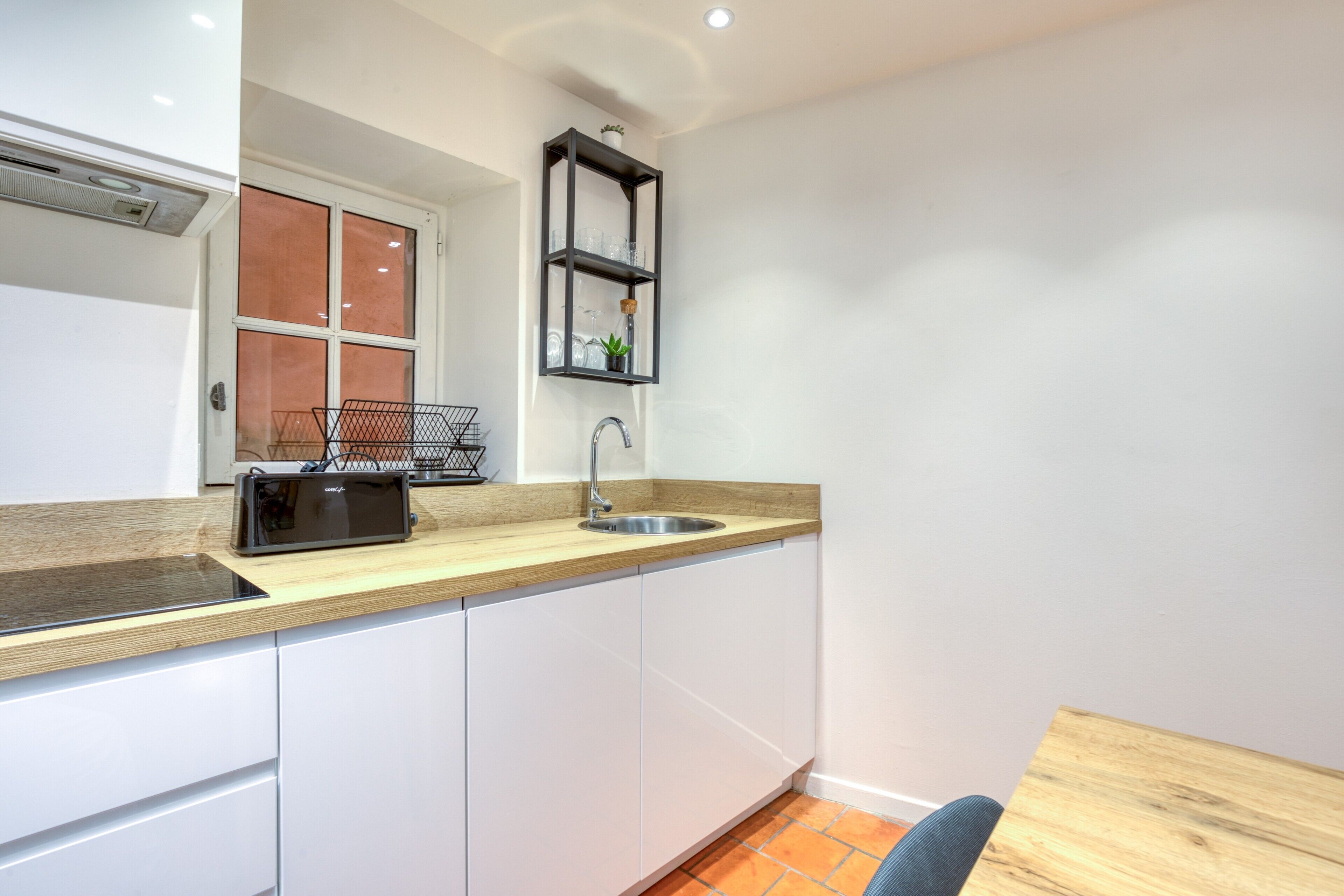 Una cocina compacta con encimera de madera, armarios blancos, una estantería negra con cristalería y una ventana con vistas.