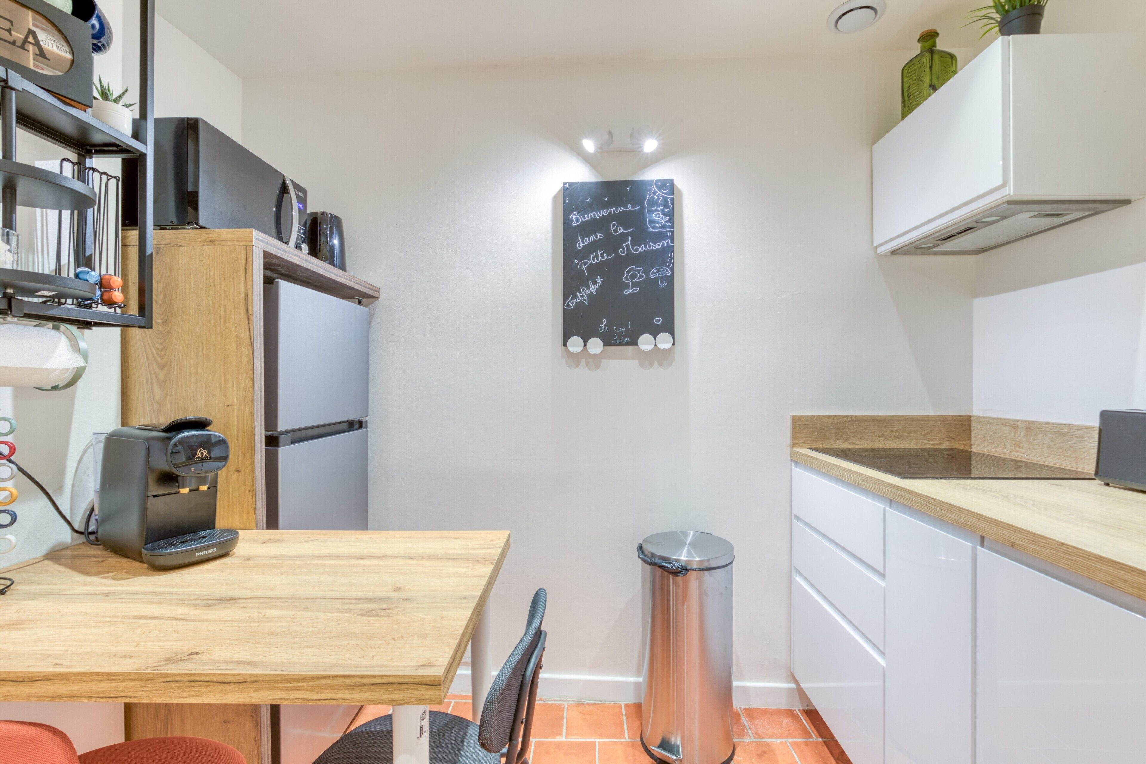 A compact kitchen with a coffee machine, a chalkboard with writing, a trash bin, and white cabinets.