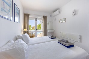 Een hotelkamer met twee bedden, een raam met gordijnen, een kleine tafel met een lamp, een wandairconditioner en uitzicht op een buitenzwembad.