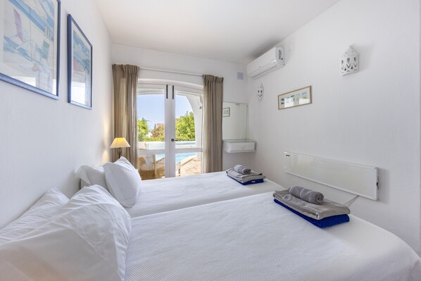 A hotel room with two beds, a window with curtains, a small table with a lamp, a wall-mounted air conditioner, and a view of an outdoor pool.