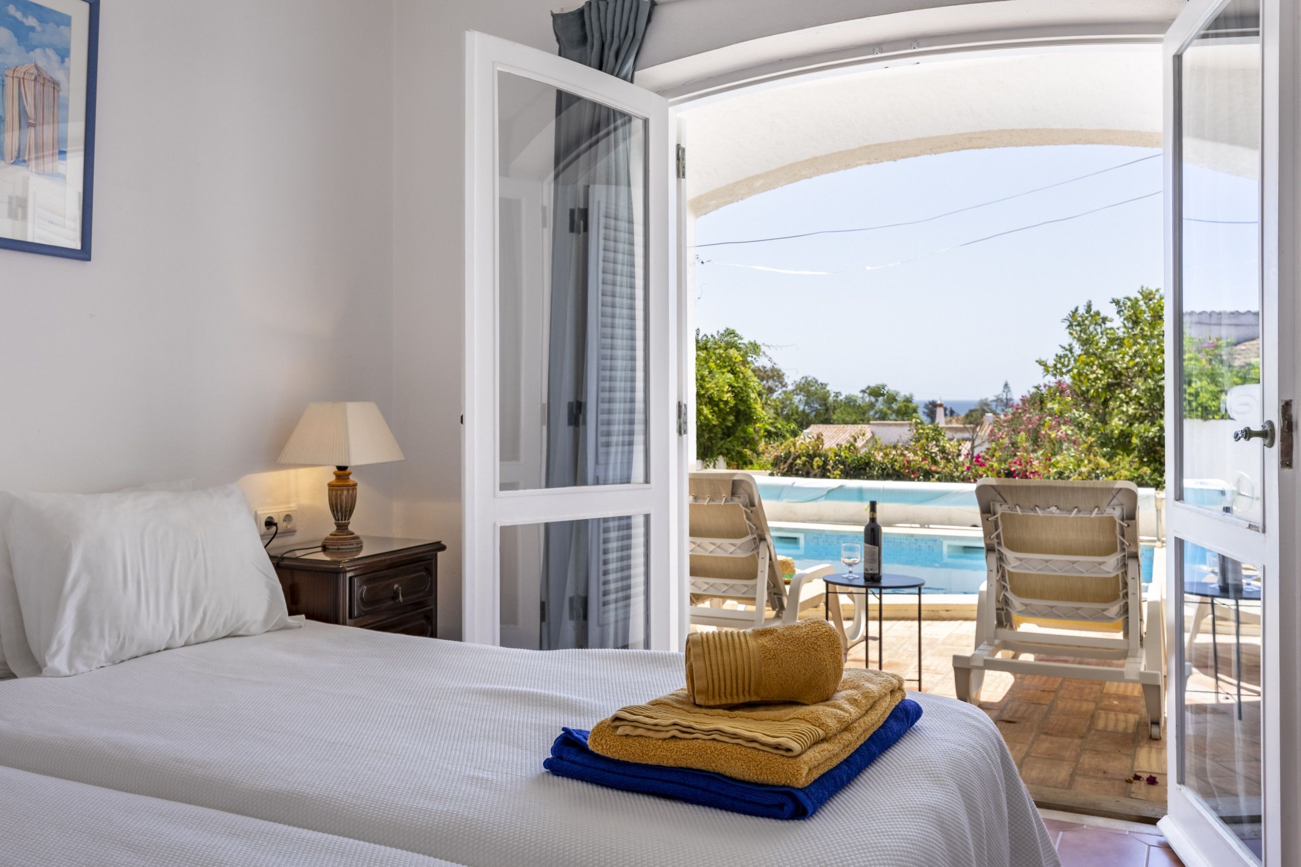 A hotel room with a bed, a nightstand, a lamp, and an open door leading to a balcony with a pool and chairs.