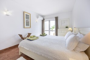 A hotel room with a bed, a chair, a table, a window, and a wall-mounted air conditioning unit. - Casa das Buganvilias 2LBC by Coolhouses (Luz)