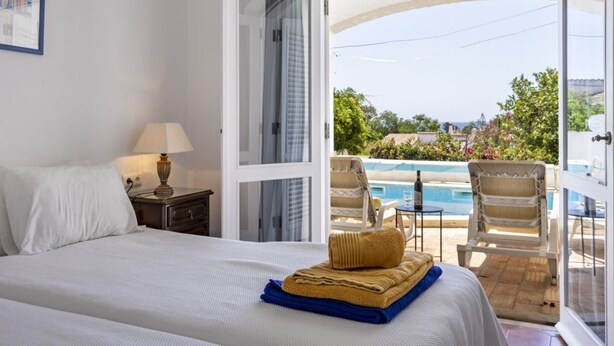 Une chambre d’hôtel comprenant un lit, une table de chevet, une lampe et donnant sur une piscine extérieure et des chaises longues.