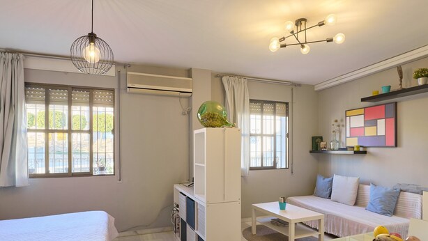 A room with a bed, a sofa, a small table, a hanging light fixture, and a window with curtains. - Casa Curro — Cozy Studio with Garden Views in Baena (Baena)