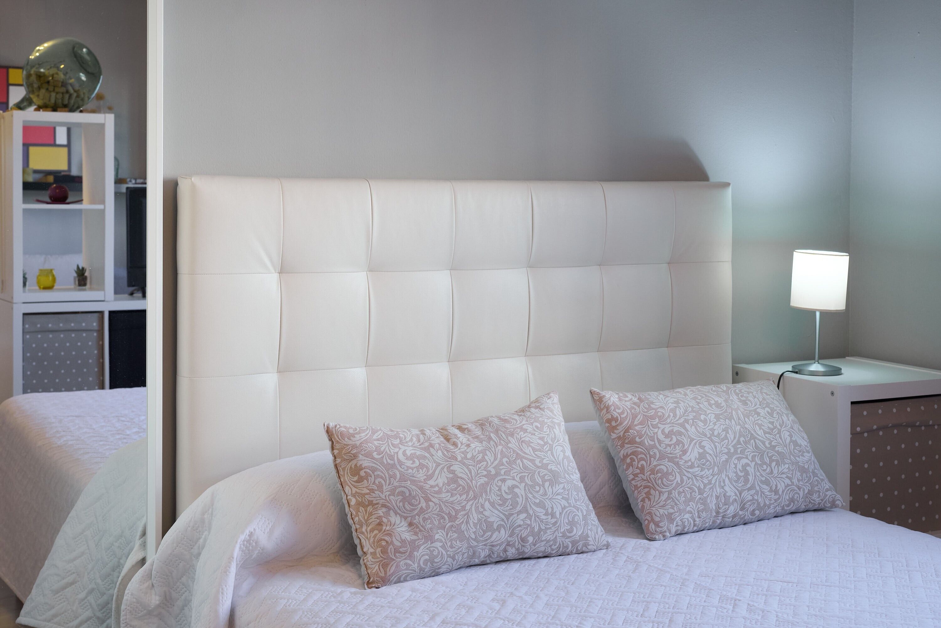 Habitación de hotel con cabecera capitonada, dos almohadas estampadas, una lámpara de noche y una mesita de noche blanca.