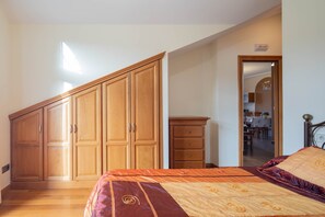 A bedroom with a large wardrobe, a bed with patterned bedding, and a view of a dining area through an open door.
