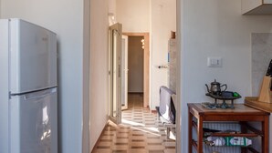 Un pasillo estrecho con un refrigerador, una puerta de madera, una silla y una pequeña mesa con utensilios de cocina.