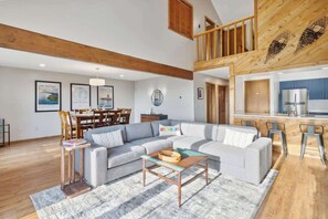 A spacious living area with a large sofa, a coffee table, and a dining table with chairs. A kitchen area with a refrigerator and cabinets is visible in the background. - Shanty Creek Retreat! (Bellaire)