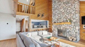 A modern living room with a stone fireplace, a sofa, a coffee table, and a kitchen area visible through an open door. - Shanty Creek Retreat! (Bellaire)