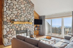 A living room with a stone wall, a fireplace, a television, and a balcony with outdoor furniture. - Shanty Creek Retreat! (Bellaire)