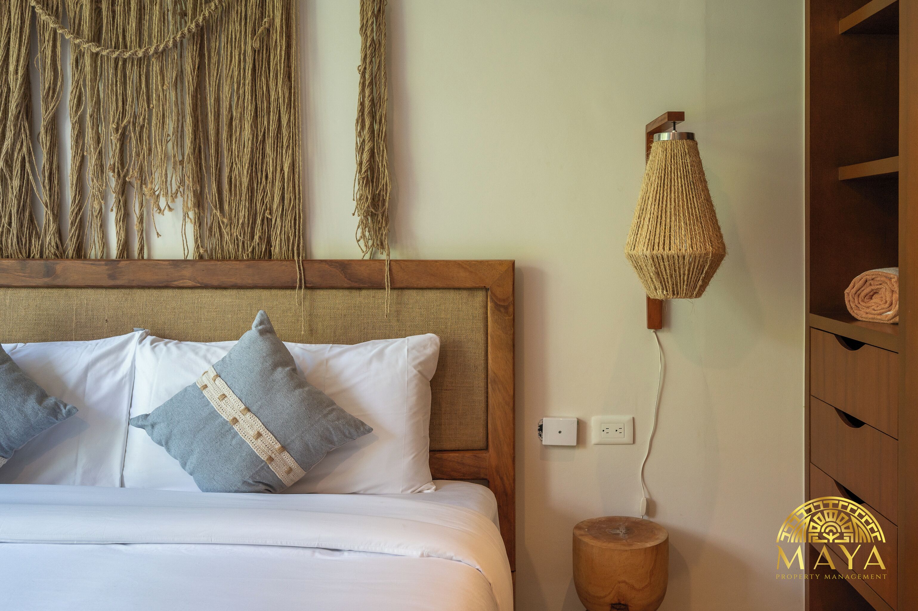 A neatly made bed with white and grey pillows, a wooden headboard, and a woven wall hanging above.