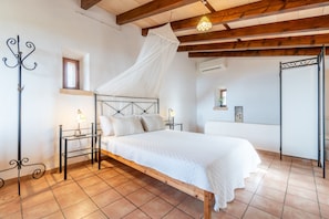 Un dormitorio con una cama de madera, ropa de cama blanca, dosel, mesita de noche con lámpara, ventana y puerta.