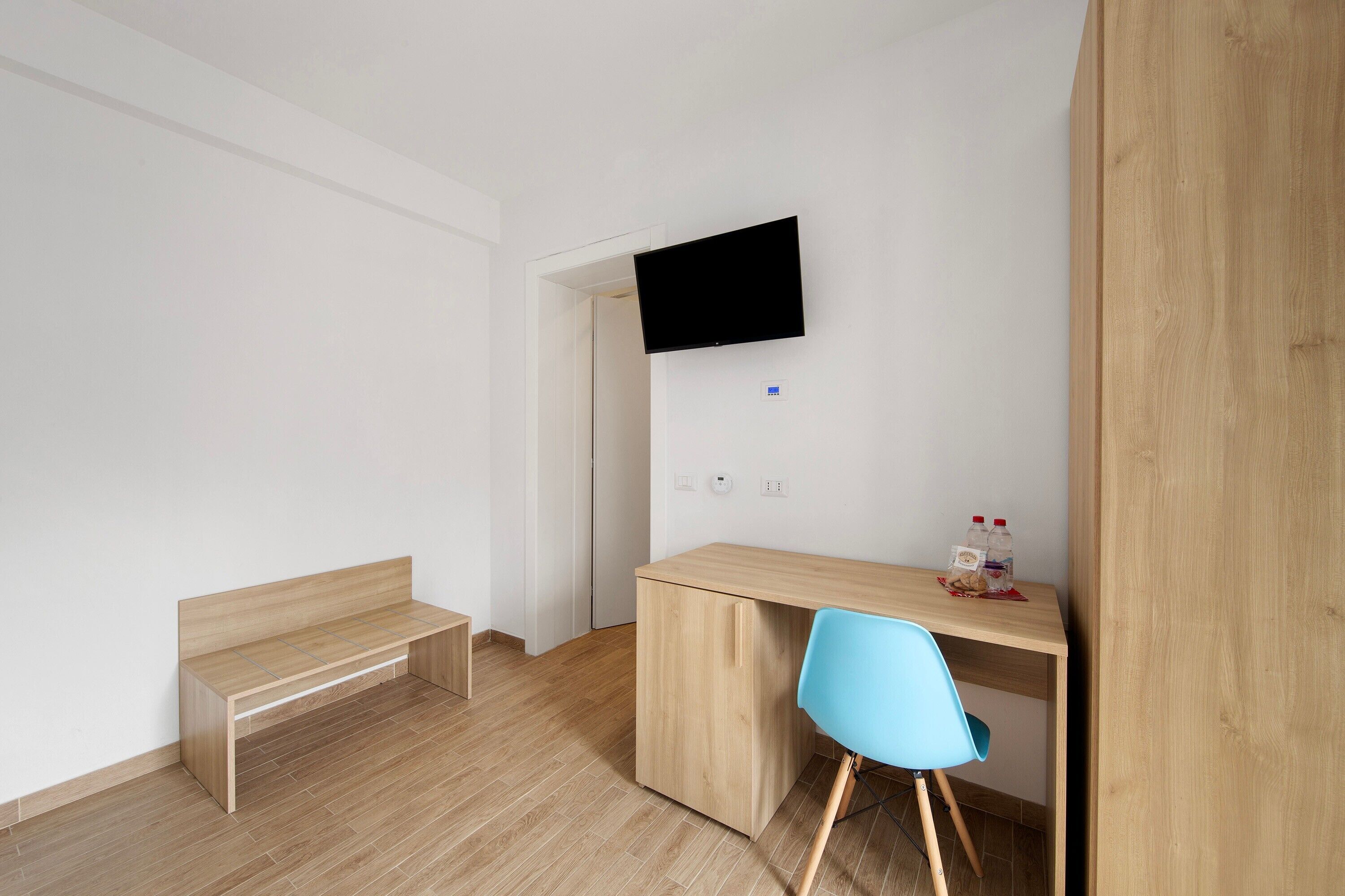 Ein Zimmer mit einem Holztisch, einem blauen Stuhl, einer Bank und einem Wandfernseher.