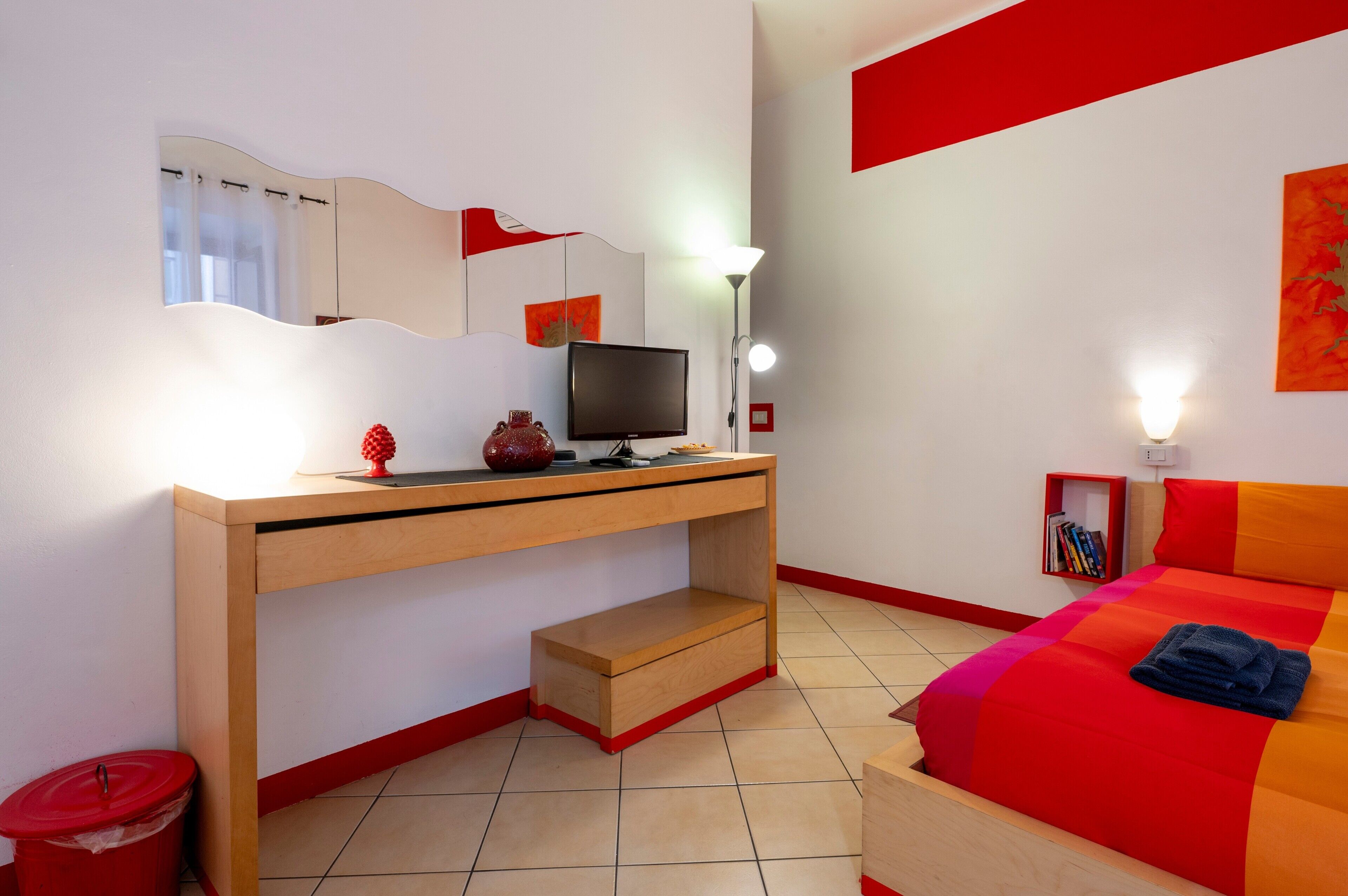 A bedroom with a bed, a desk with a computer, a lamp, a mirror, and a bookshelf.