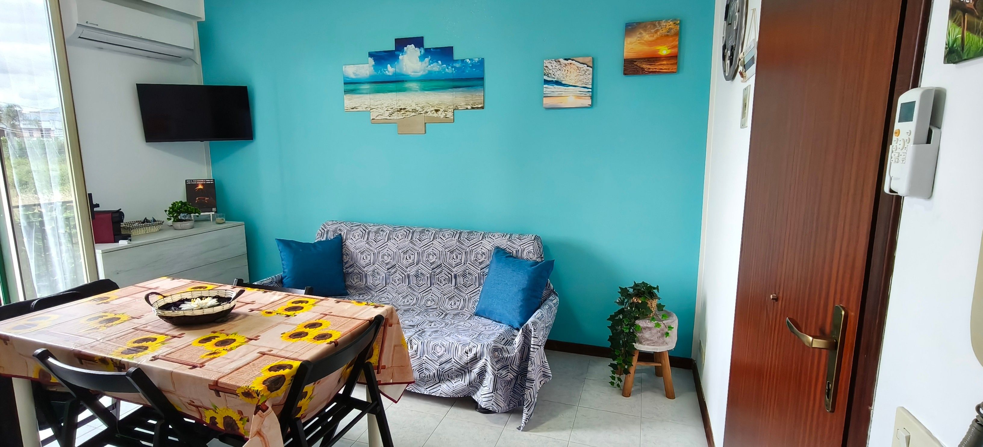 A room with a teal wall, a patterned sofa, a dining table with a sunflower tablecloth, and a small potted plant.
