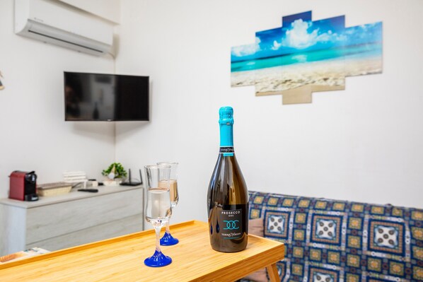 A bottle of Prosecco on a tray with two glasses, a TV, and a beach-themed wall art in a room with patterned cushions.