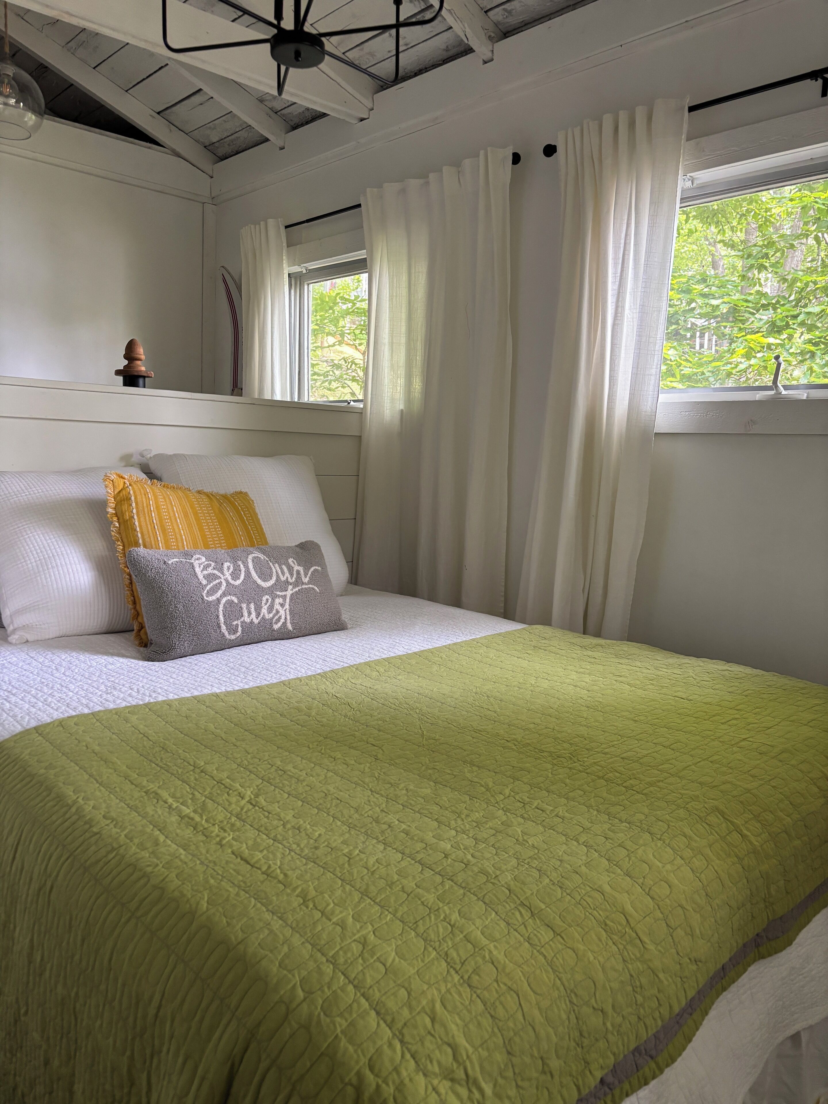 Un lit bien fait, avec une couette verte, des oreillers blancs et un coussin orné de la phrase « Soyez les bienvenus ».